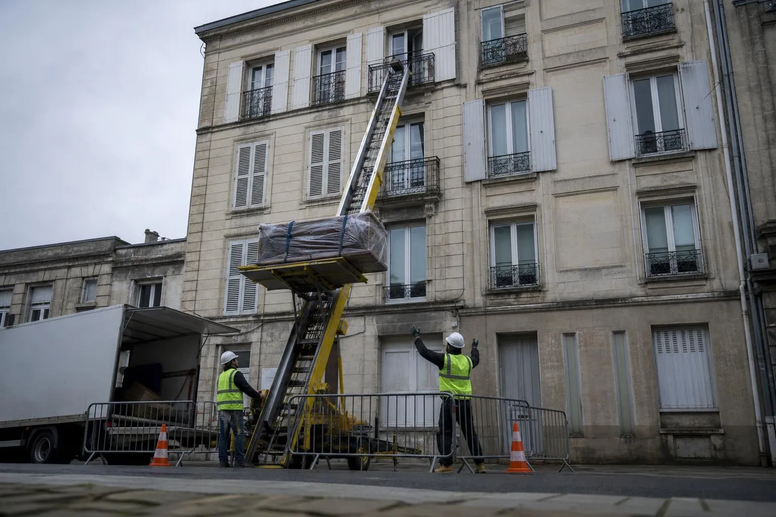Un monte meuble déployé le long d’une façade d’immeuble hisse un canapé emballé vers le troisième étage, deux hommes le guident depuis le trottoir